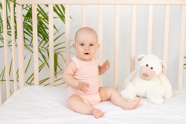 Baby girl in a crib in a pink bodysuit and a teddy bear six months