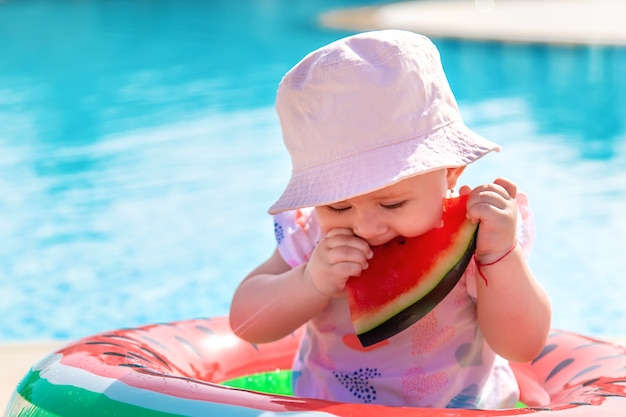 Baby girl eats watermelon in a watermelon inflatable by the swimming pool