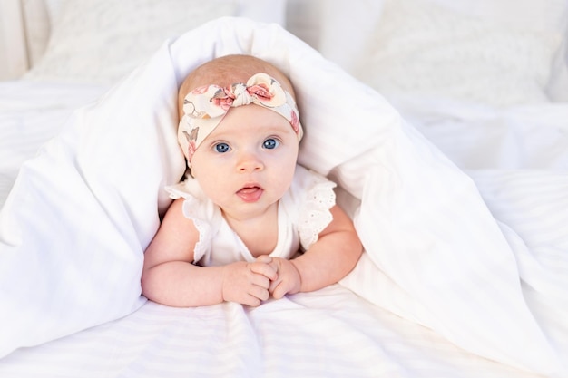 Baby girl lies under a blanket and smiles on a white cotton bed at home newborn morning