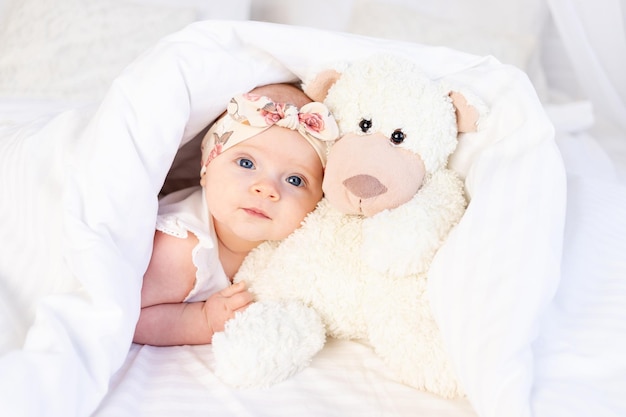 A baby girl lies under a blanket with a teddy bear toy and smiles on a white cotton bed at home