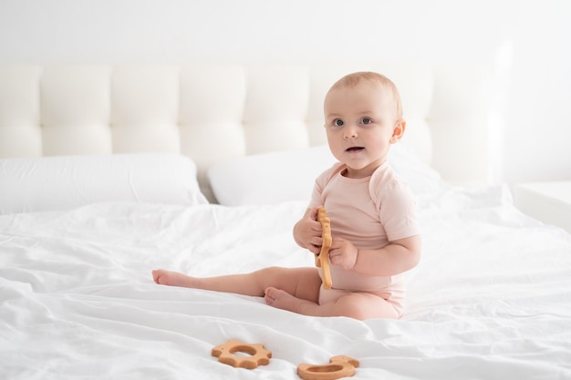 Baby girl in light pink bodysuit playing with wooden toys on white bedding on bed