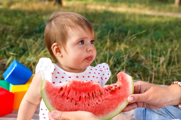 baby girl in the park in the summer with her mother tries watermelon for the first time