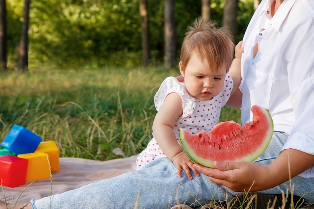 baby girl in the park in the summer with her mother tries watermelon for the first time