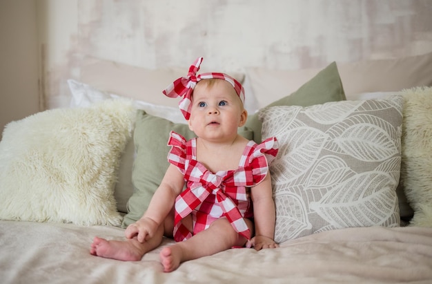 A baby girl in a plaid bodysuit is sitting on a bed in a room