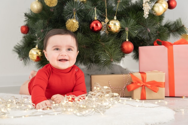 Baby girl in red bodysuit under Christmas tree with gift boxes presents