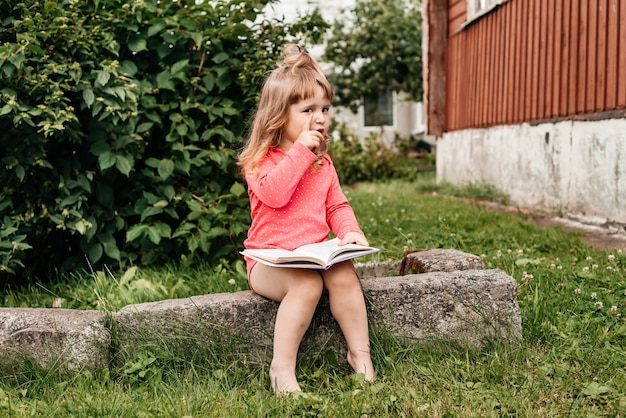 Baby girl in a red on green grass and reading a book in a sunny summer day.