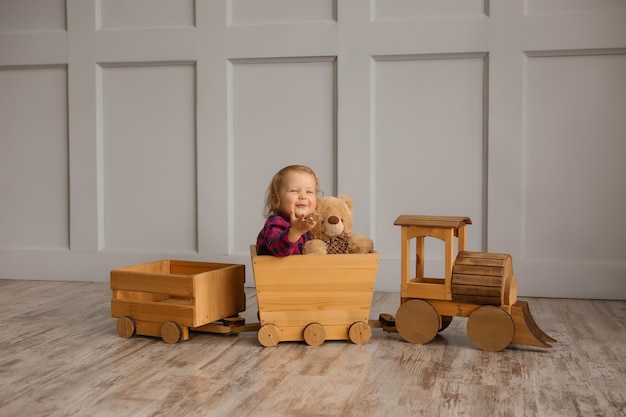 Baby girl smiling holding a Teddy bear, sitting in a toy wooden locomotive