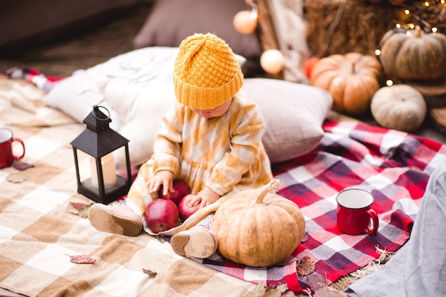 Baby girl wearing knitted hat and stylish dress holding red tasty apples with pumpkin