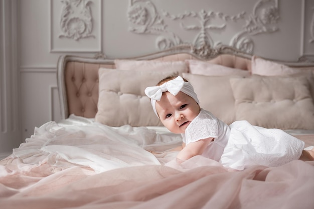 A baby girl in a white headband and bodysuit crawls on a bed in a room with a beautiful interior