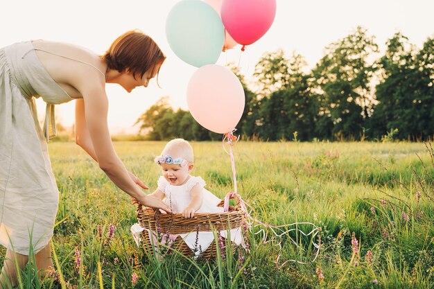 Baby girl in wicker basket with balloons in sunlight at sunset Mother and child outdoors First birthday Family celebrates one year old baby on nature Photo of healthy dreams vacation holidays