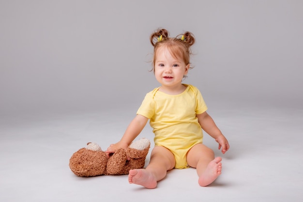 A baby girl in a yellow bodysuit sits and plays with a teddy bear on a white background