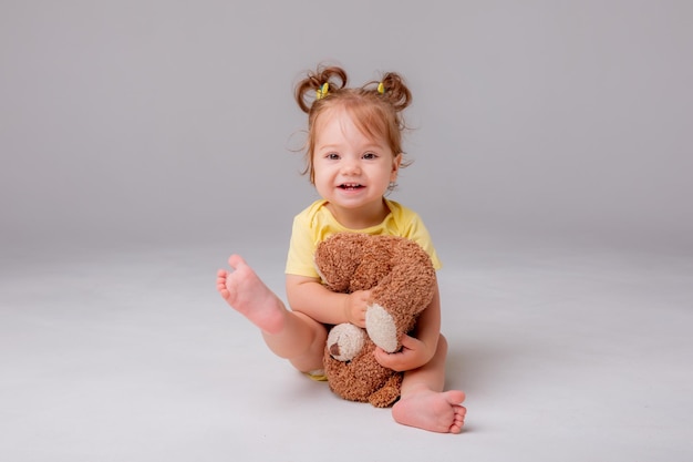 A baby girl in a yellow bodysuit sits and plays with a teddy bear on a white background