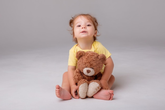 A baby girl in a yellow bodysuit sits and plays with a teddy bear on a white background