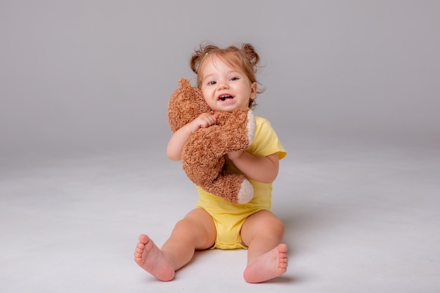 A baby girl in a yellow bodysuit sits and plays with a teddy bear on a white background