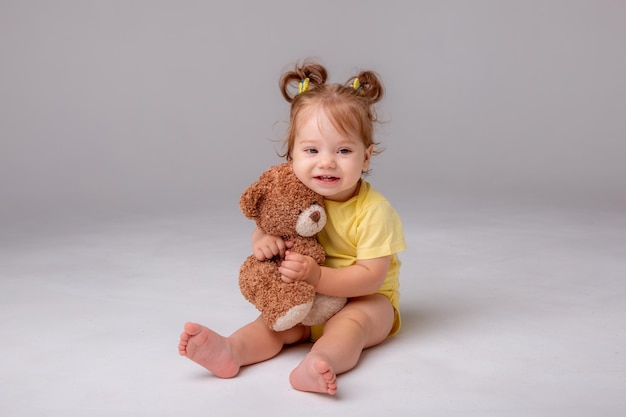 A baby girl in a yellow bodysuit sits and plays with a teddy bear on a white background