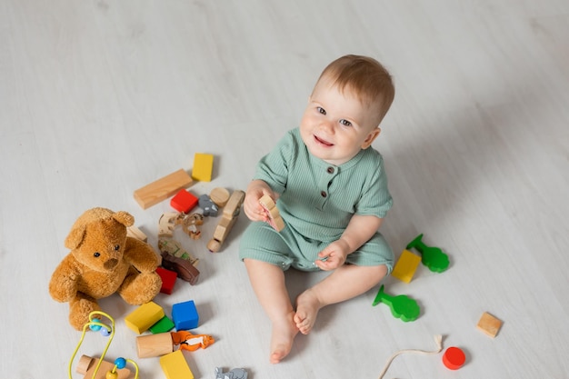 Baby in a green bodysuit is sitting on the wooden floor of the house playing educational wooden toys