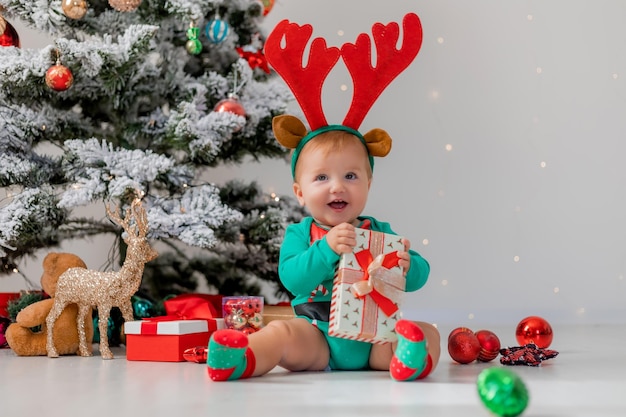 Baby in a green bodysuit with red deer horns on his head is playing with gift boxes near Christmas tree. child in a Christmas gnome costume. winter New Year's concept. holiday goods. space for text