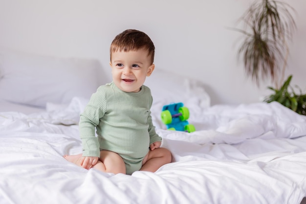 Baby in a green cotton bodysuit is sitting on a white bed with a colored toy car child is playing