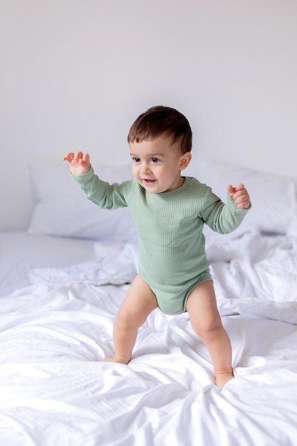 Baby in a green cotton bodysuit stands barefoot on a white bed