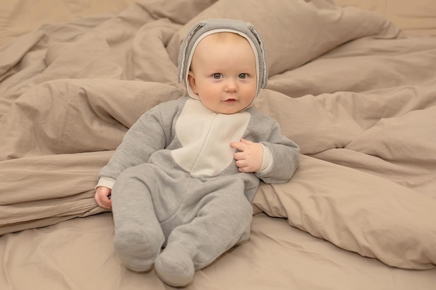 A baby in a grey animal suit sits on a bed.