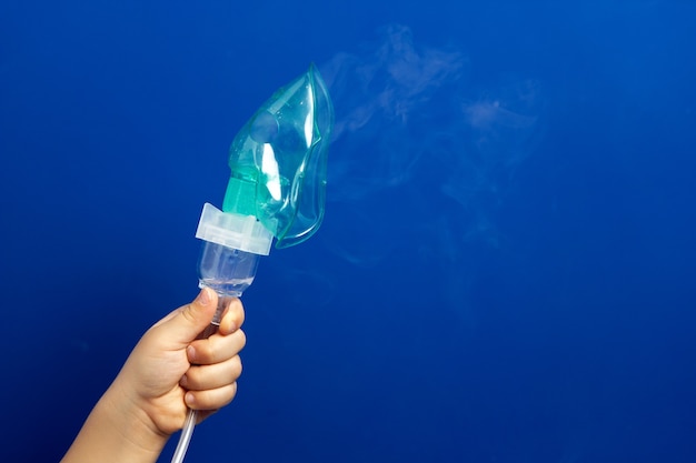 Baby hand holding green rubber mask for breathing treatment on a blue wall