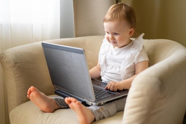 A baby in a light chair sits at a laptop and spoils his eyesight watching cartoons