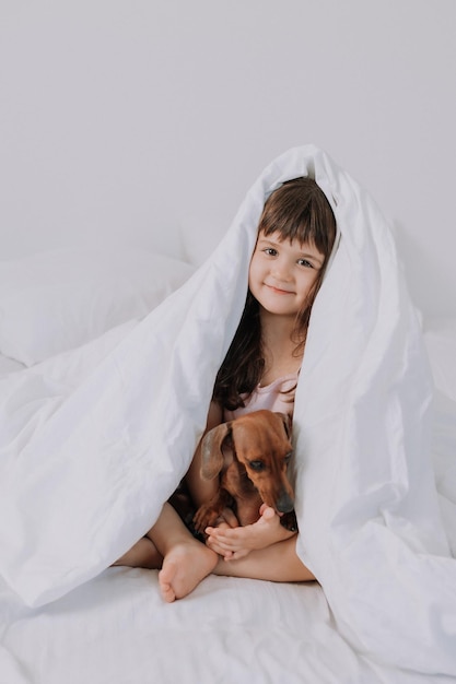 baby little girl hugs dachshund dog at home in bed. white bed linen. love for pets