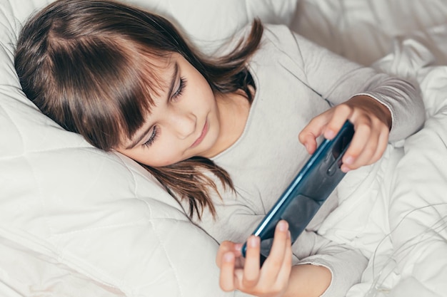 Baby little girl lying in bed with phone and watching cartoons or playing a game on the phone and smiling