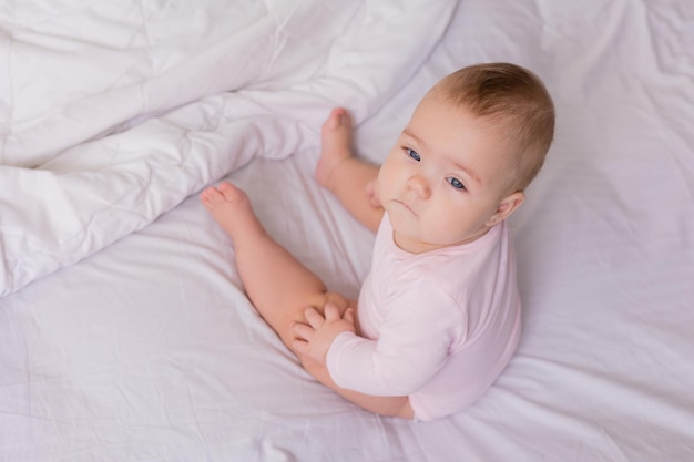 baby in a pink bodysuit is sitting with her back to the camera in a crib on snowwhite bed linen