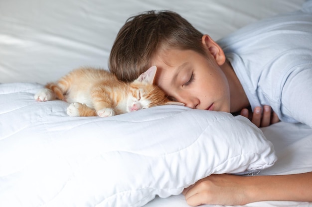 Baby and red cat sleeping together on white sheet kitten and kid childhood friendship concept Kid and fluffy pet