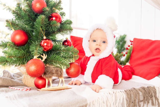 A baby in a Santa Claus costume under the Christmas tree plays with red Christmas balls lying on the window of the house the concept of the new year