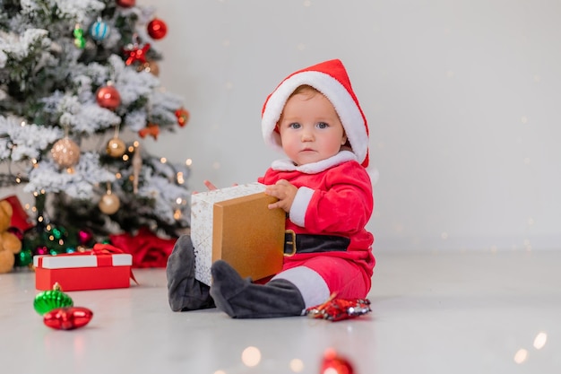 Baby in a Santa costume is sitting next to the Christmas tree and playing with gift boxes. child opens a Christmas gift. winter new year's concept. space for text. High quality photo