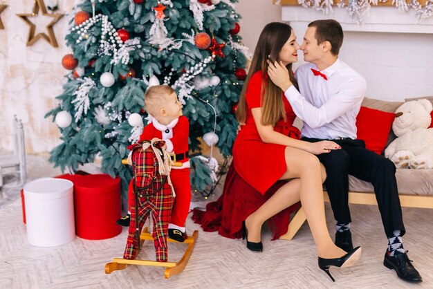 baby sits on rocking horse near christmas tree and gift mom and dad are sitting on the couch and kissing