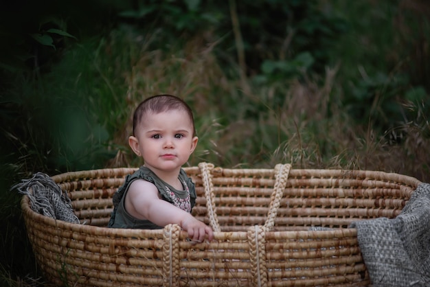 The baby sits in a straw wicker cradle in a green park. Little girl in cute dress smiling. Portrait.