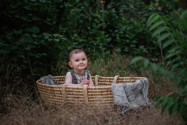 The baby sits in a straw wicker cradle in a green park. Little girl in cute dress smiling. Portrait.