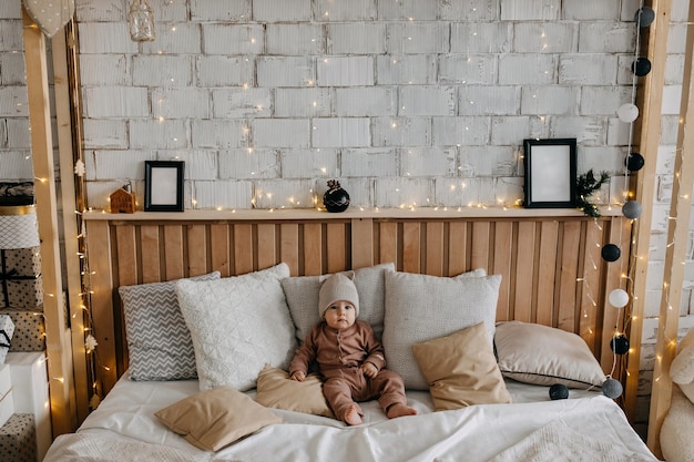 Baby sitting on bed between pillows at home barefoot wearing a beanie with ears