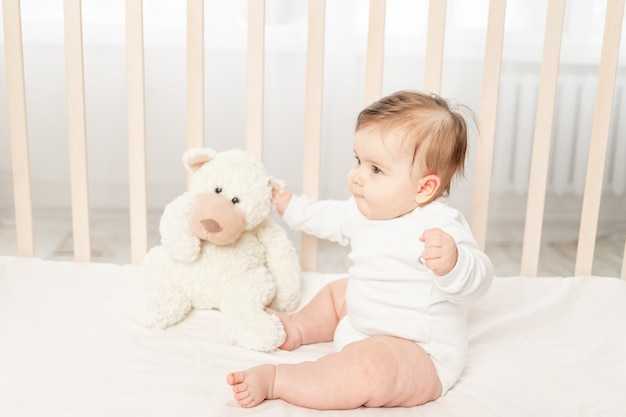 Baby six months playing in a crib in a white bodysuit with a Teddy bear