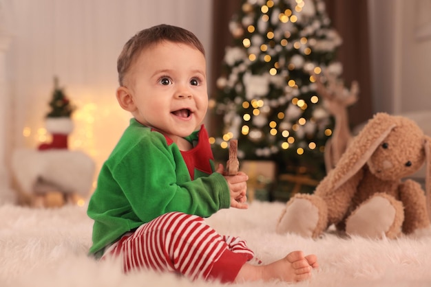 Baby wearing cute elf costume on floor in room decorated for Christmas