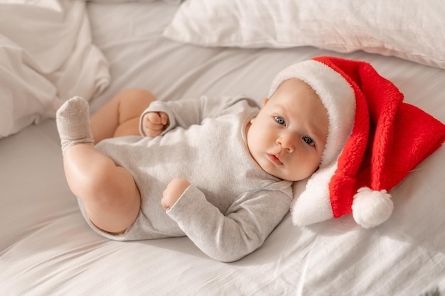 Baby in a white bodysuit and a Santa hat is lying in bed next to the Christmas tree. charming blue-eyed child. winter holiday, funny moments, child christmas. space for text. High quality photo