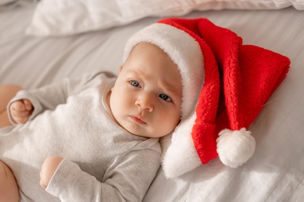 Baby in a white bodysuit and a Santa hat is lying in bed next to the Christmas tree