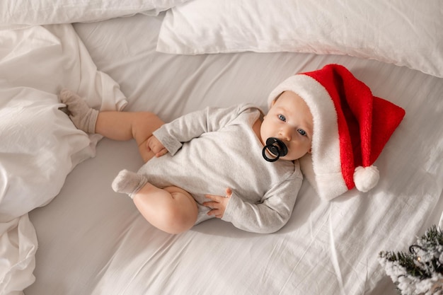 Baby in a white bodysuit and a Santa hat with black pacifier in his mouth is lying in bed top view