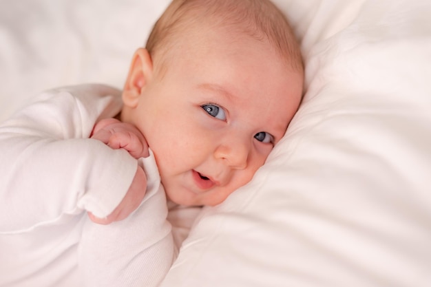 Baby with blue eyes in a white bodysuit lies on the back pressing his arms to his chest on white bed