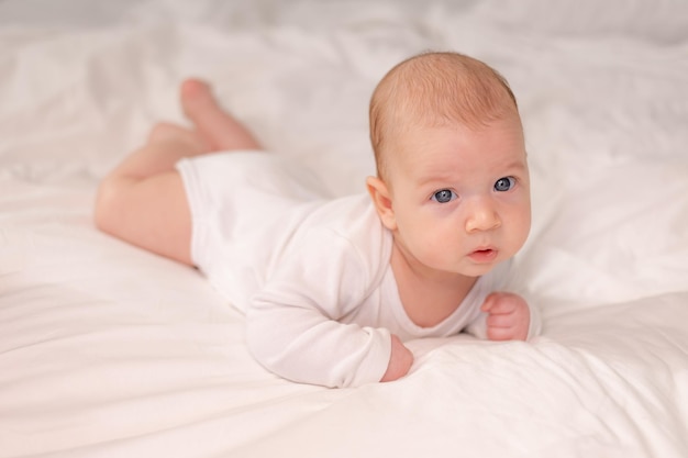 Baby with blue eyes in a white bodysuit lies on his tummy on white bed linen lifestyle space for