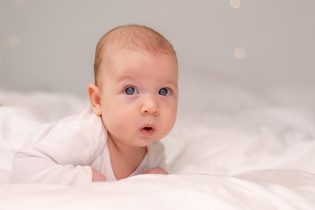 Baby with blue eyes in a white bodysuit lies on his tummy on white bed linen lifestyle