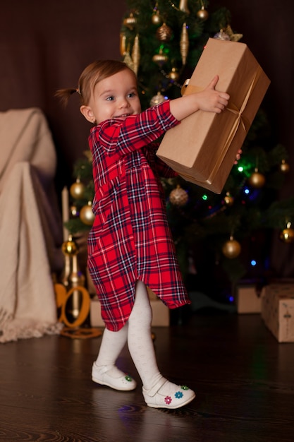 Baby with light brown hair in a red checkered dress holding a big Christmasgift dark color