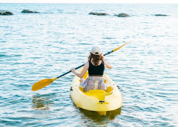 Back of Asian woman in black tank top and cap kayaking on yellow kayak boat with using paddle on the sea Happy female having fun activity on seascape view background Holiday trip vacation