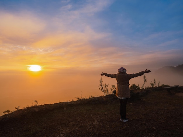 Back of Asian woman with freedom arms open standing on mountain peak at Pha Tang view point 104 with river view and sea mist and sun rise in fog sky. travel place in Chaing Rai, Thailand