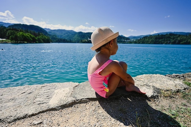 Back of baby girl is swimsuit and hat sit in pier of view beautiful Bled Lake Slovenia