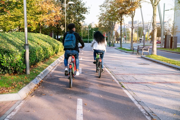 Back camera two young men and a woman riding a bike path with a shared electric bike in a beautiful park with many trees at sunset