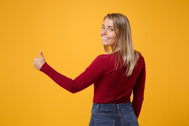 Back rear view of smiling young blonde woman girl in casual clothes posing isolated on yellow orange wall background. People lifestyle concept. Mock up copy space. Showing thumb up looking camera.